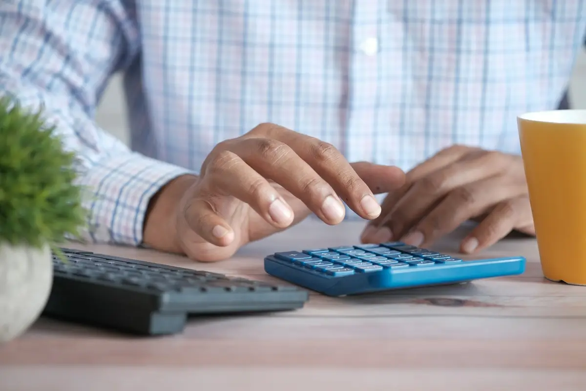 Mãos digitam numa calculadora azul sobre a mesa, ao lado de um teclado