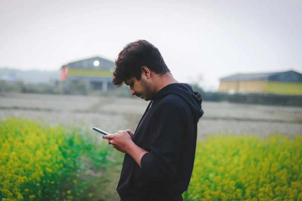 Homem olha a tela do celular em pé numa área de plantação