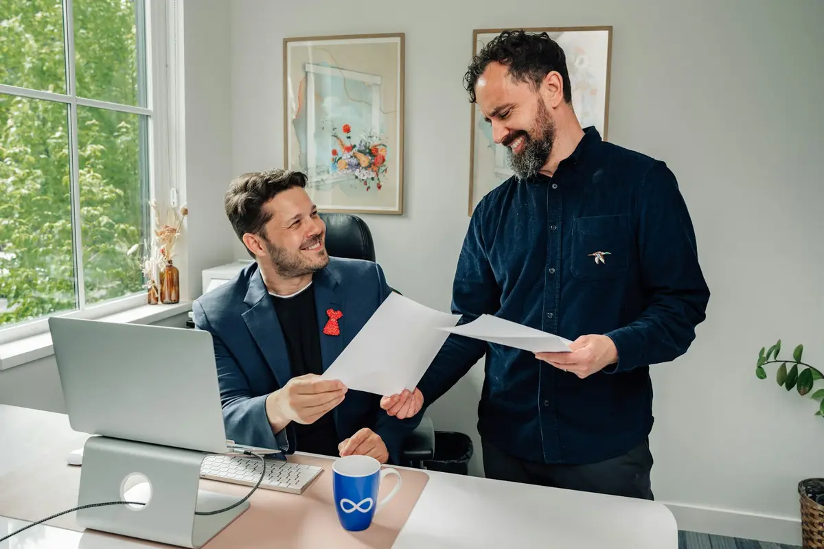 Dois homens conferem folhas de papel numa mesa de escritório com notebook
